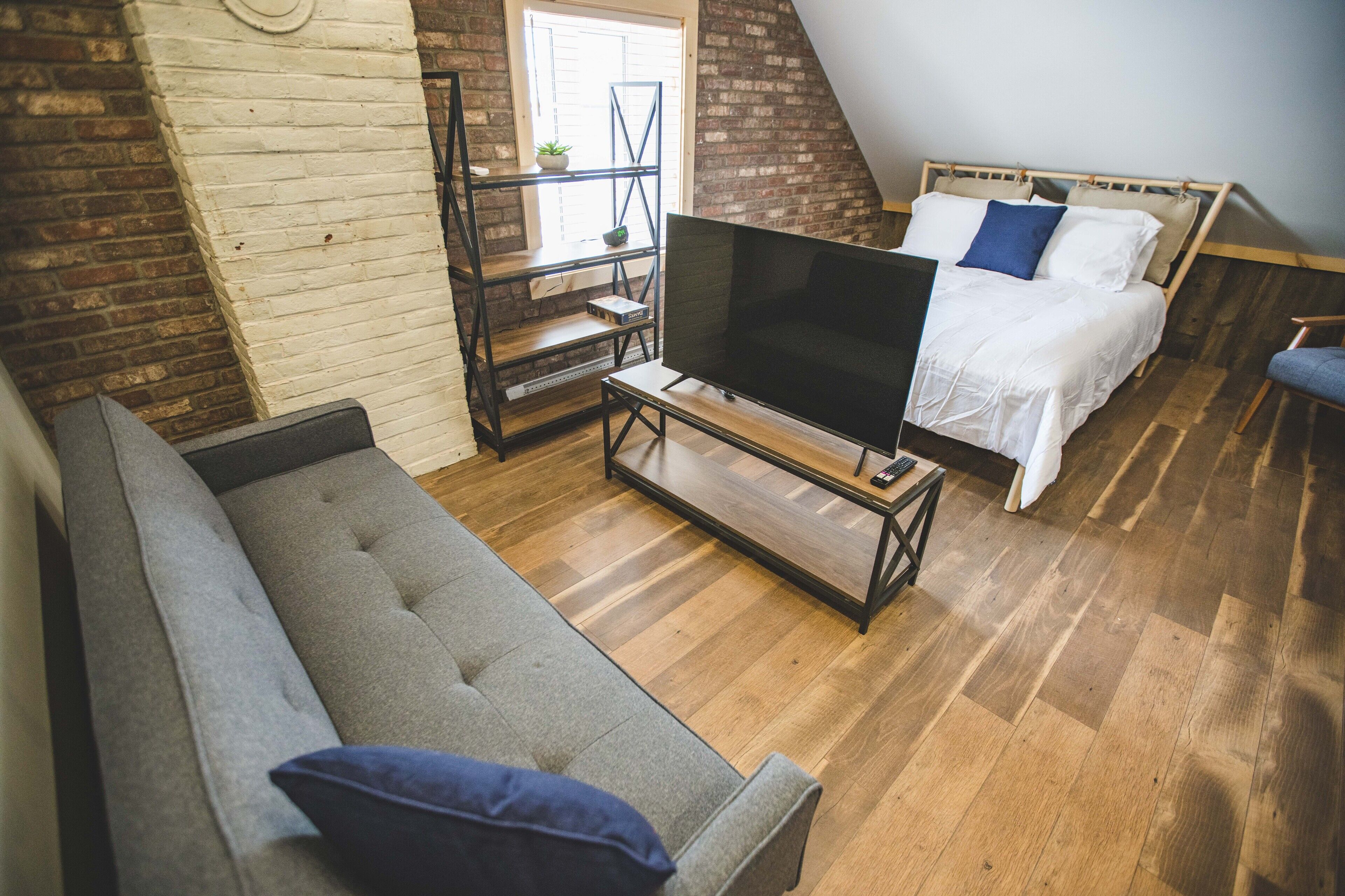 A loft-style bedroom with a wooden floor, a gray sofa, a bed with white bedding, and a TV on a wooden stand.