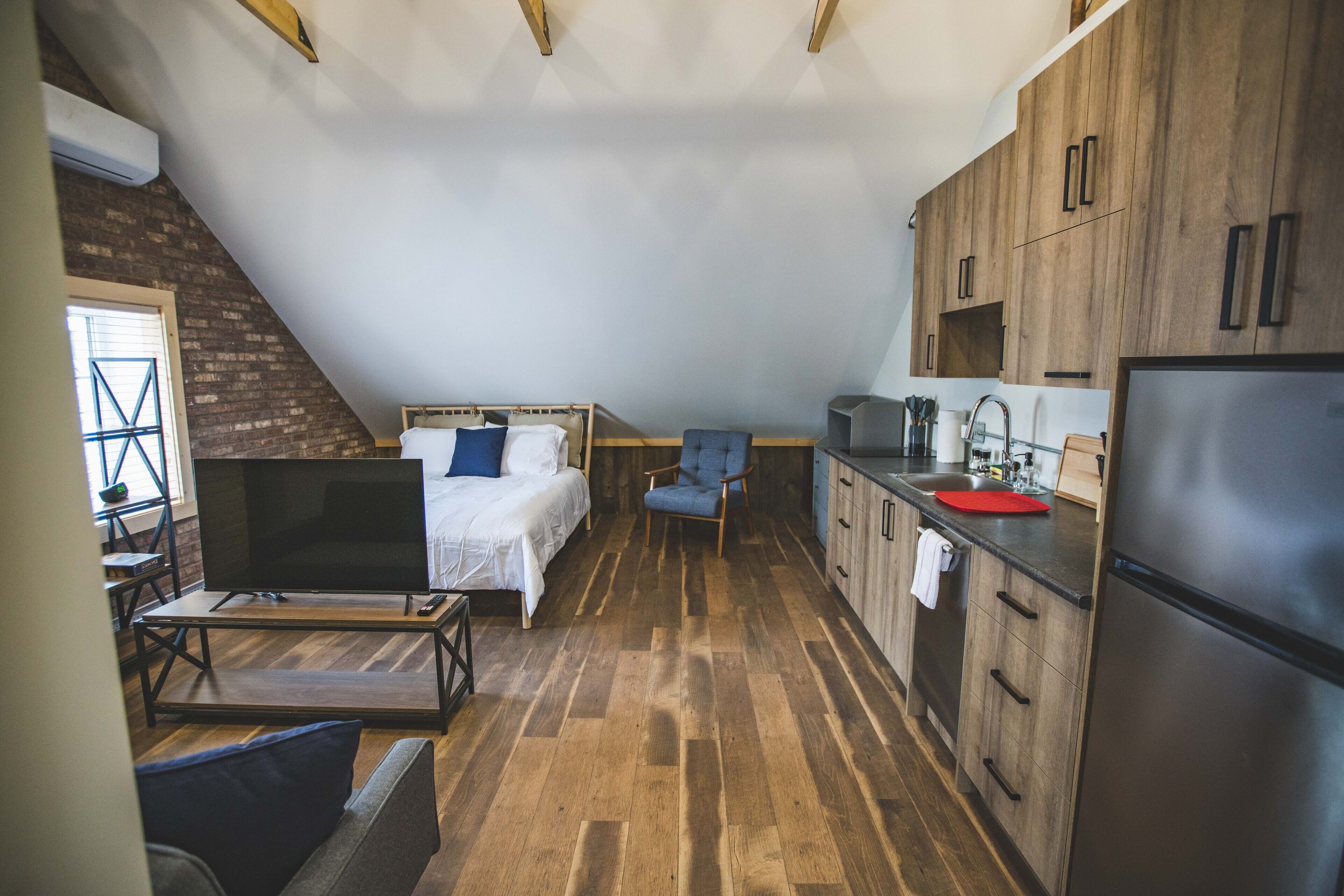 A compact room with a bed, a TV, a chair, and a kitchenette.