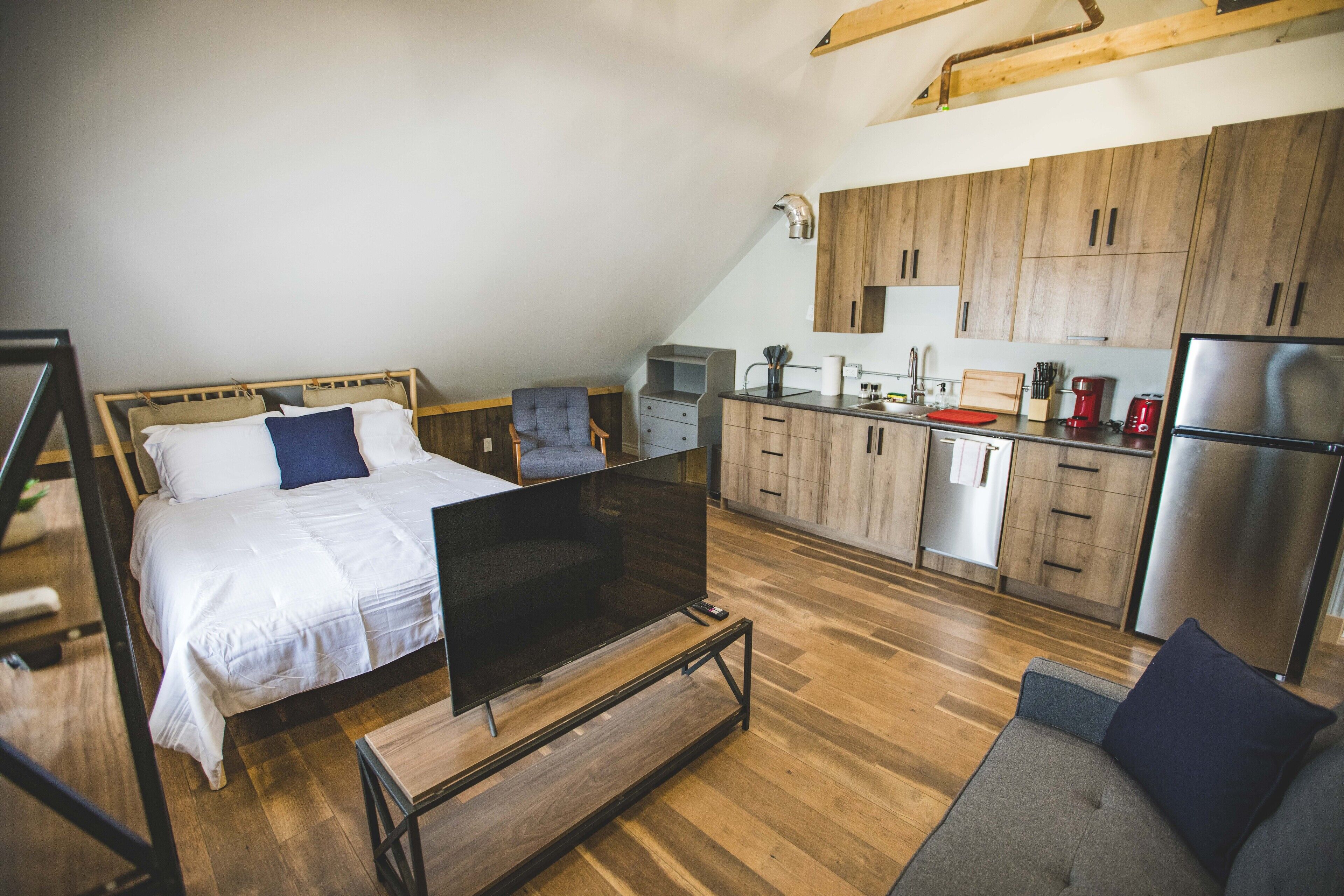 A modern bedroom with a bed, a TV, and a sofa. The room has wooden flooring and a kitchen with wooden cabinets and appliances.