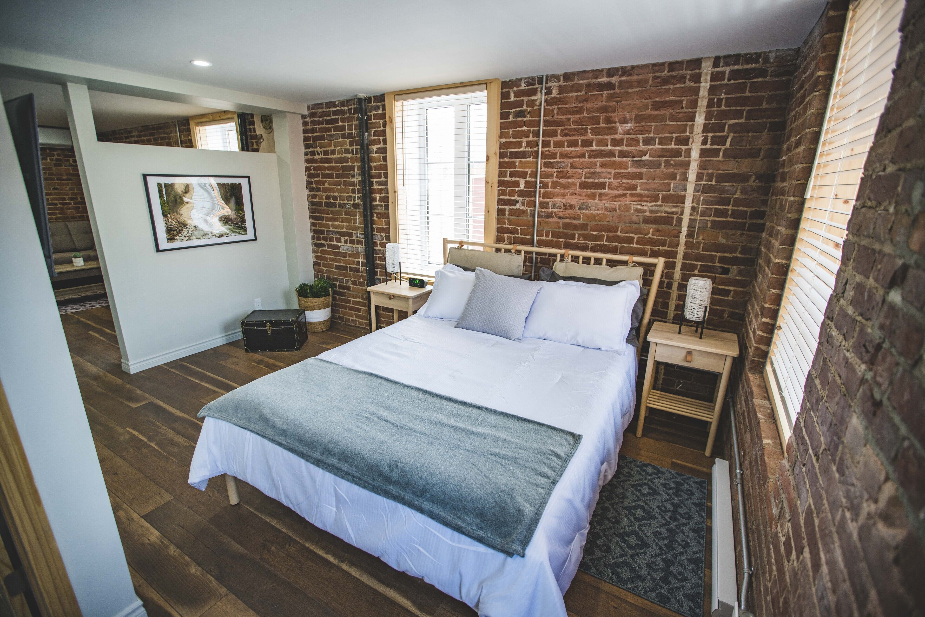 A bedroom with a large bed, a wooden headboard, a nightstand, a bedside table, a window with blinds, and a brick wall.