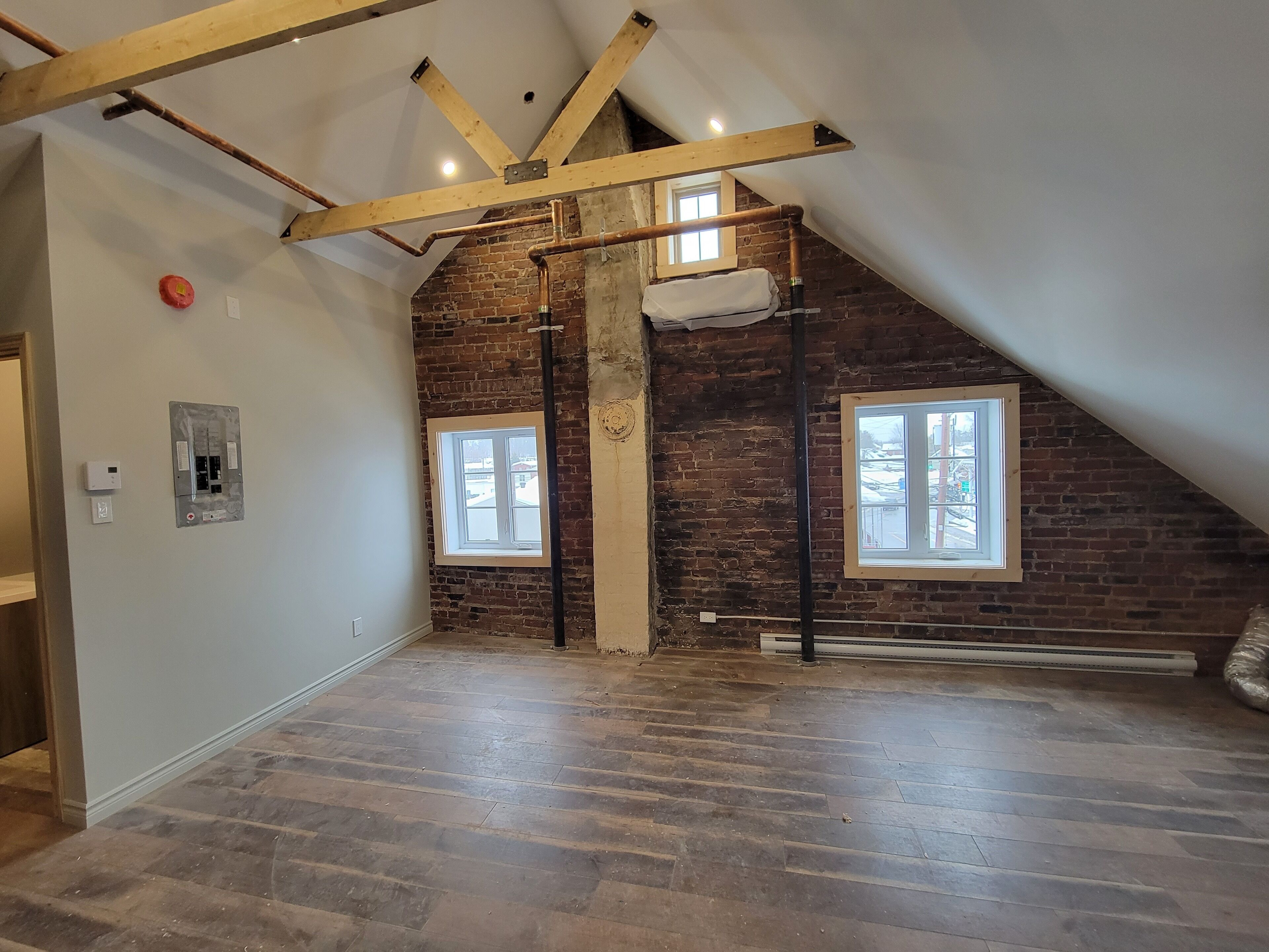 An attic room with exposed wooden beams, brick walls, and two windows.