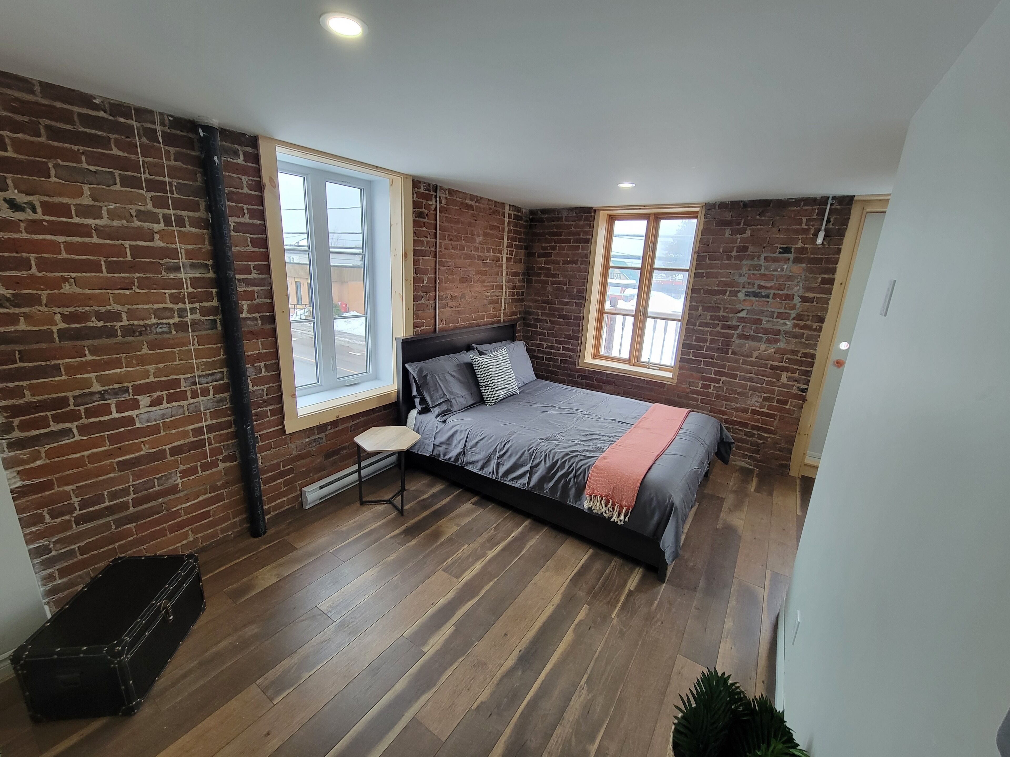 A bedroom with a brick wall, a bed, two windows, and a small table.