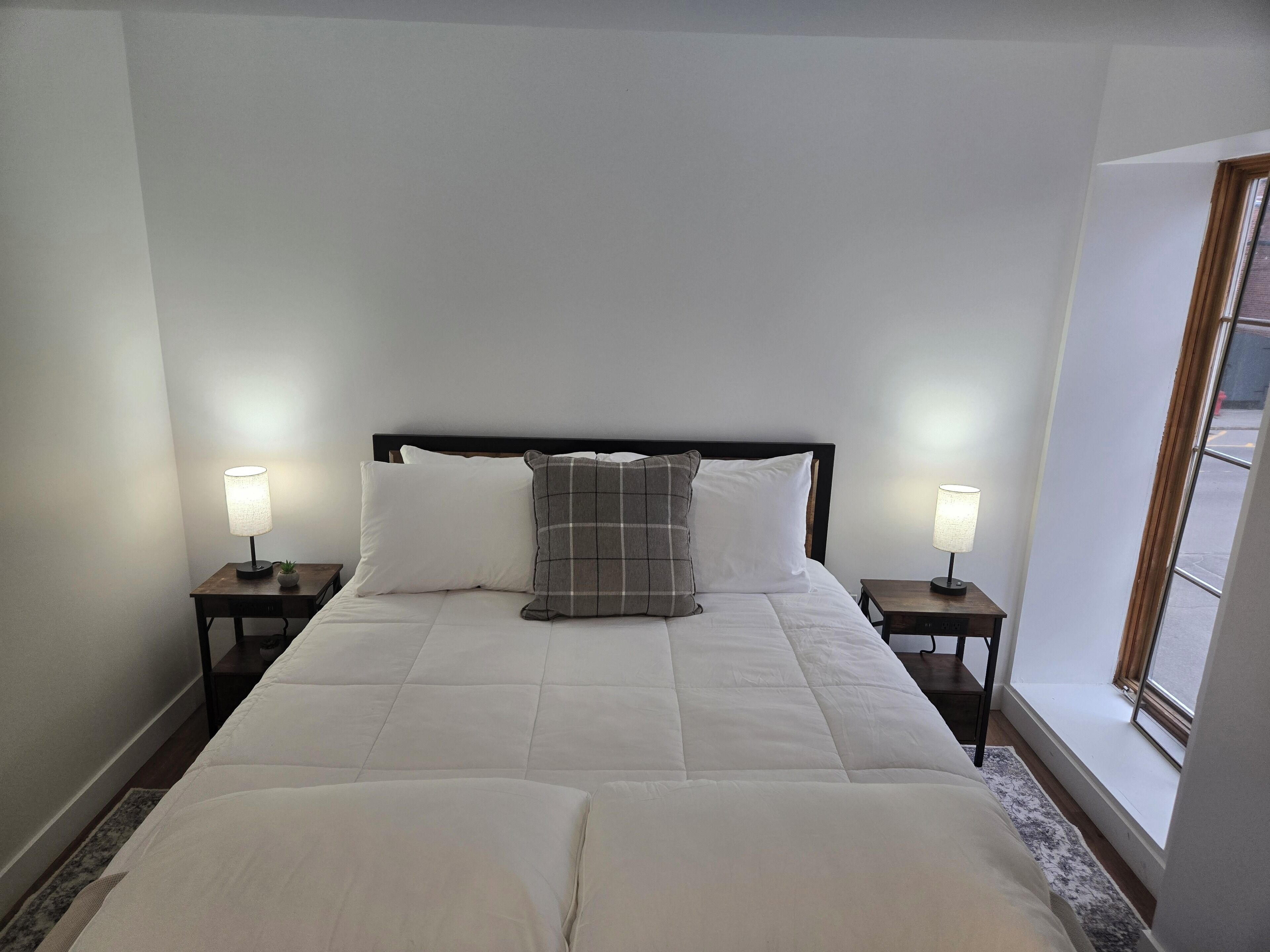 Una cama bien hecha con ropa blanca y un cojín a cuadros grises, flanqueada por dos mesitas de noche con lámparas, en una habitación con un ventanal grande.