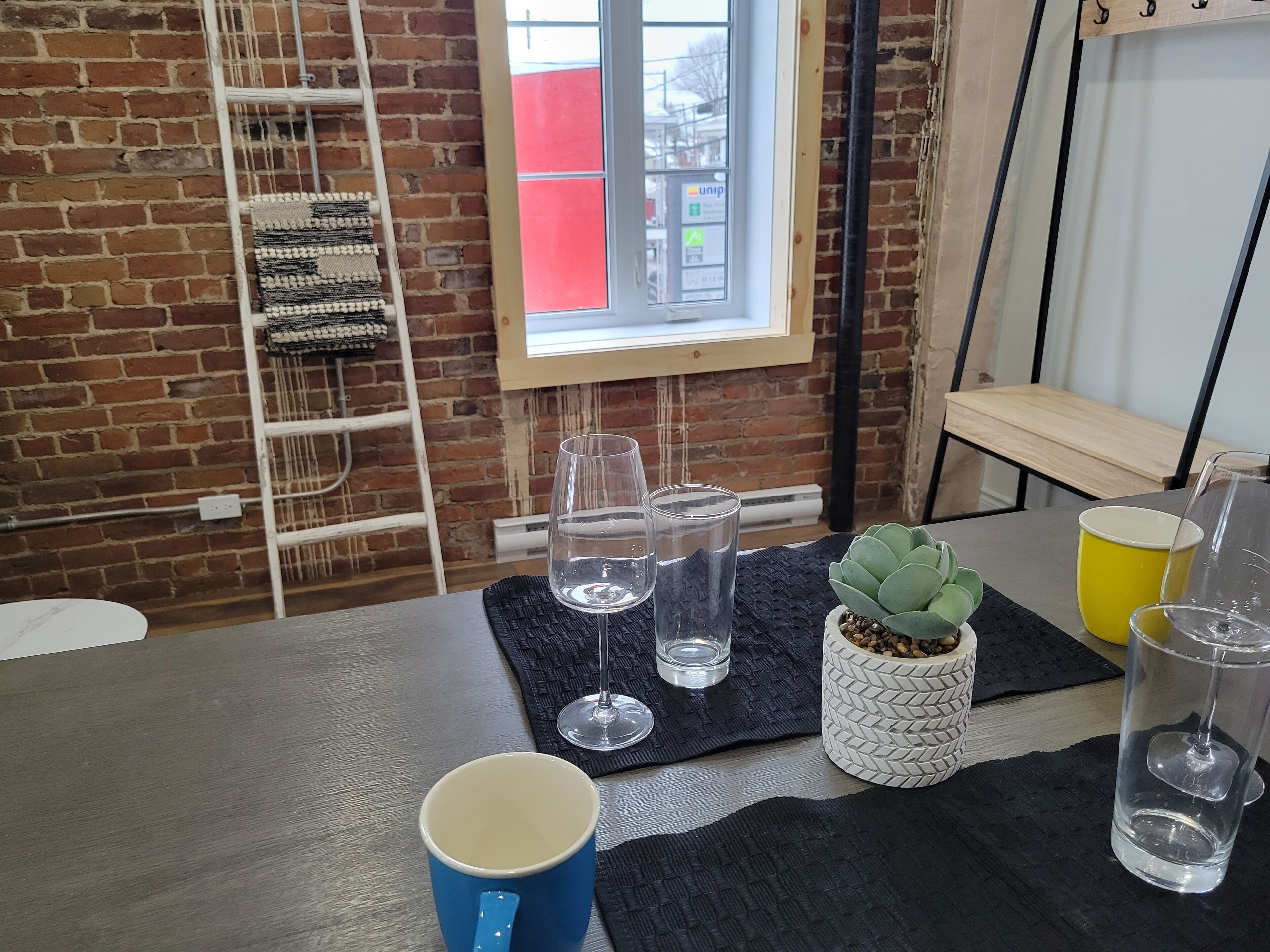 A room with a brick wall, a window with a view, a table with a blue mug, clear glasses, and a potted plant.