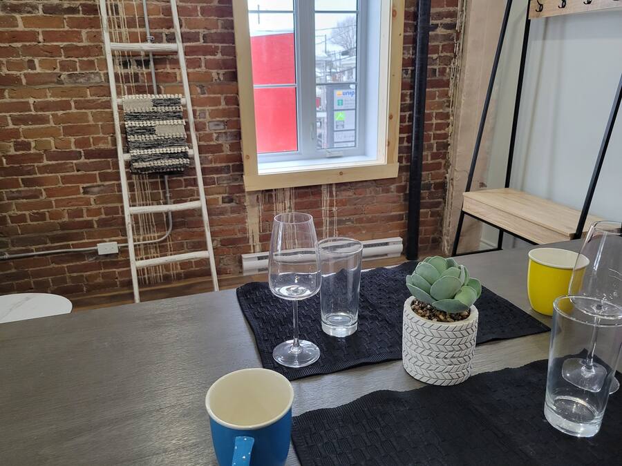 A room with a brick wall, a window with a view, a table with a blue mug, clear glasses, and a potted plant.