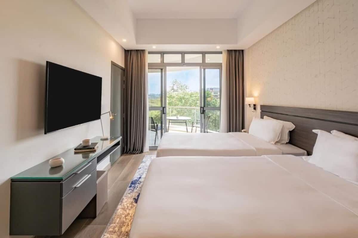 Une chambre d’hôtel avec deux lits, un téléviseur à écran plat, un bureau avec une lampe et un balcon offrant une vue sur la ville.
