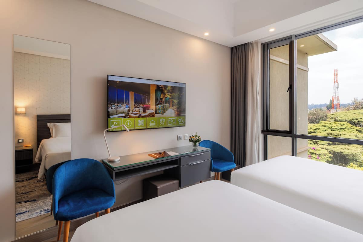 Une chambre d’hôtel moderne dotée d’une grande télévision, d’un bureau, de deux fauteuils bleus, d’un lit et offrant une vue sur la campagne.