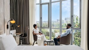 A modern hotel room with a large bed, a desk with a chair, a lamp, and a view of the city through large windows.