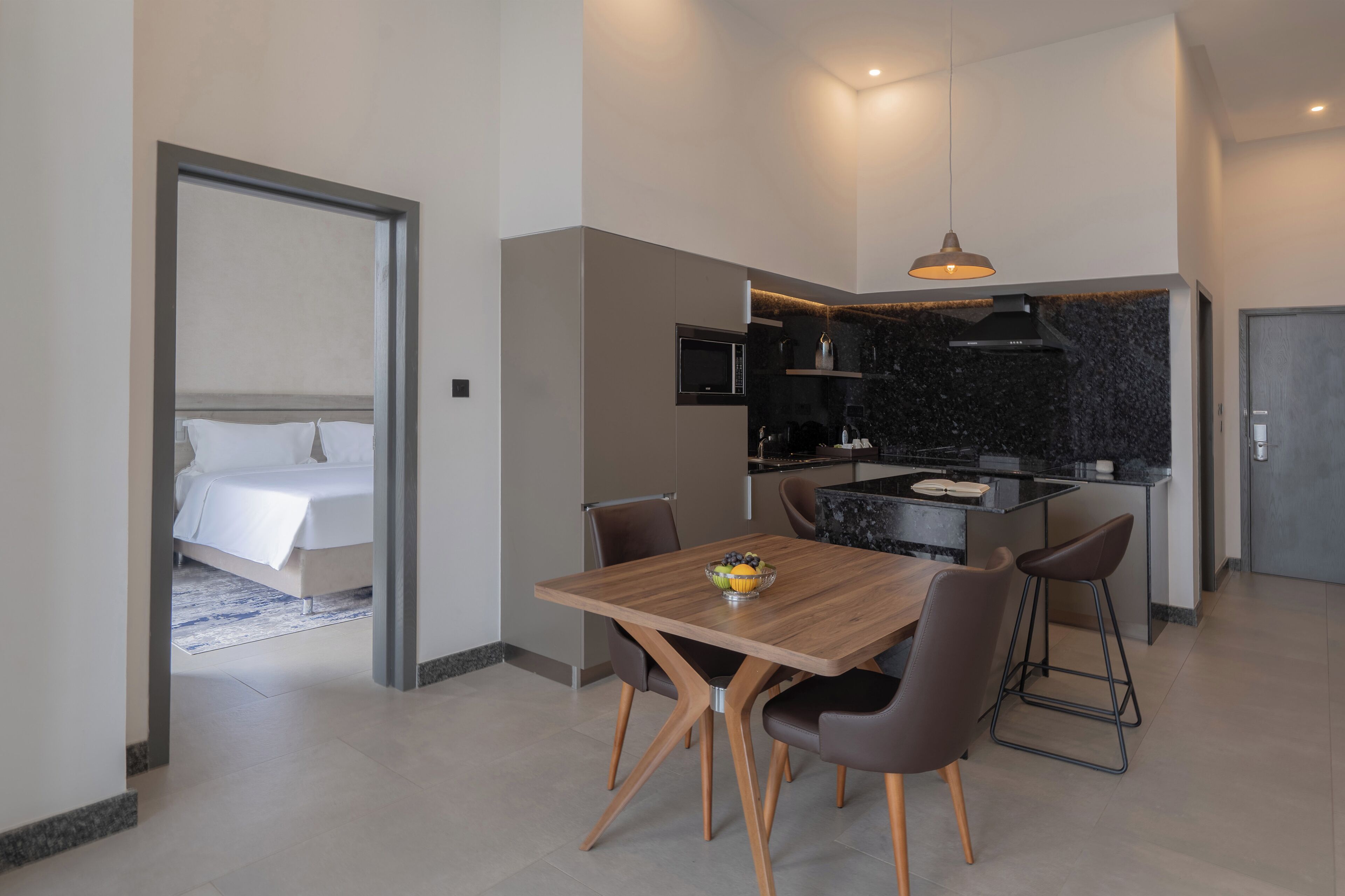 Et moderne hotelværelse med et træbord, to barstole, et køkken med en sort bordplade, og en seng i det tilstødende rum.