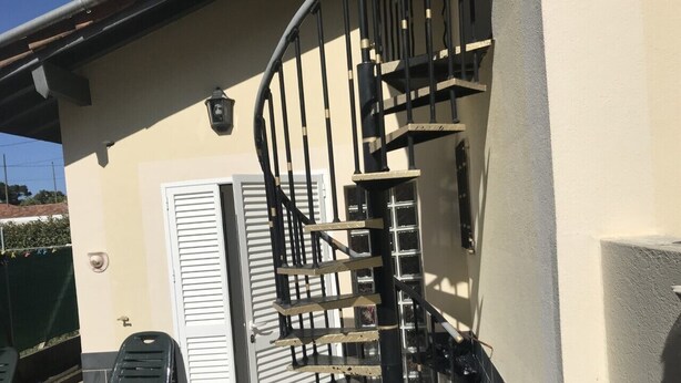 Una escalera de caracol que conduce a una puerta, zona de estar al aire libre con sillas, y un edificio con fachada color crema.