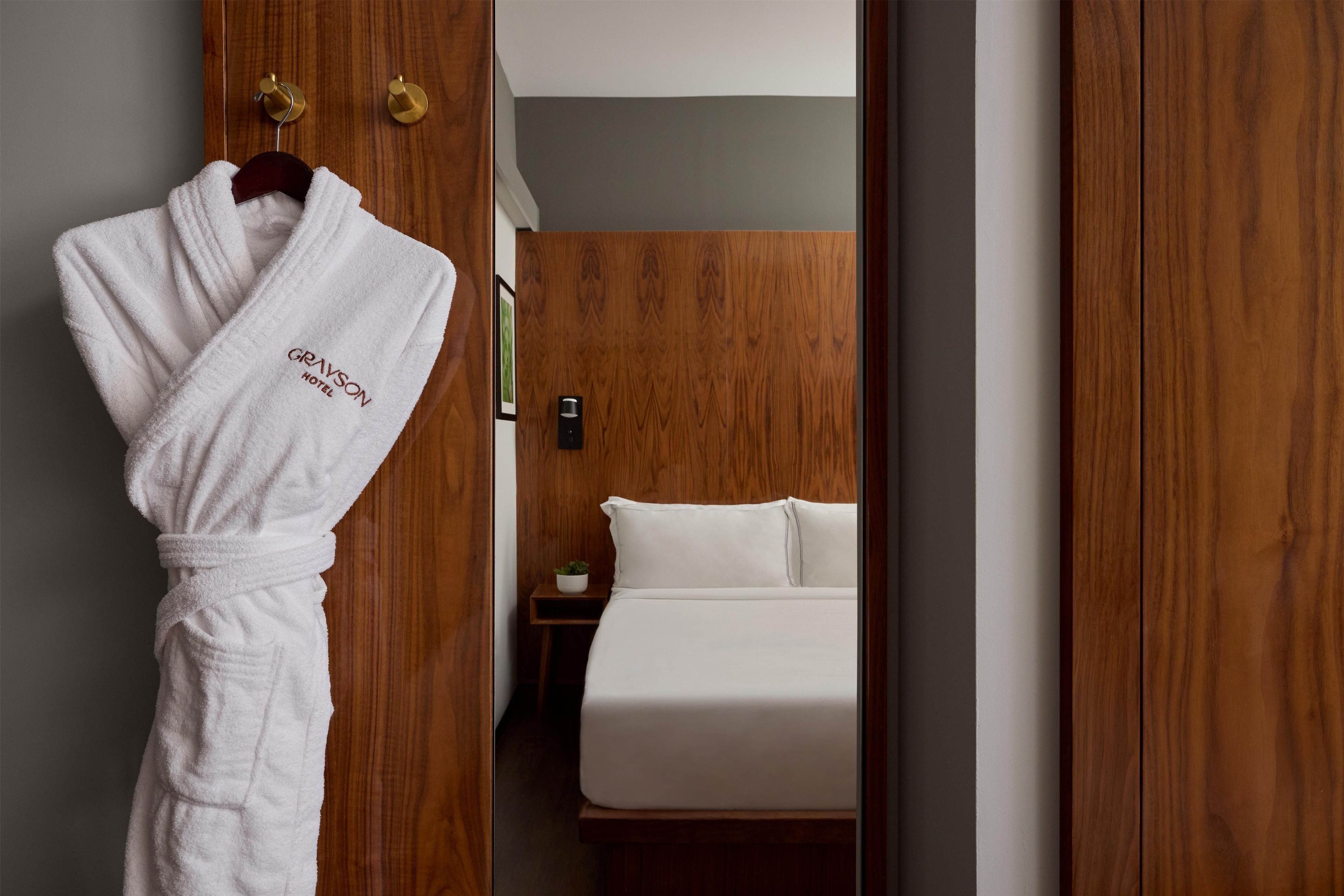 Hotelkamer met een houten garderobe, een witte badjas hangend aan de deur, een bed met wit beddengoed en een klein nachtkastje met een plantje.