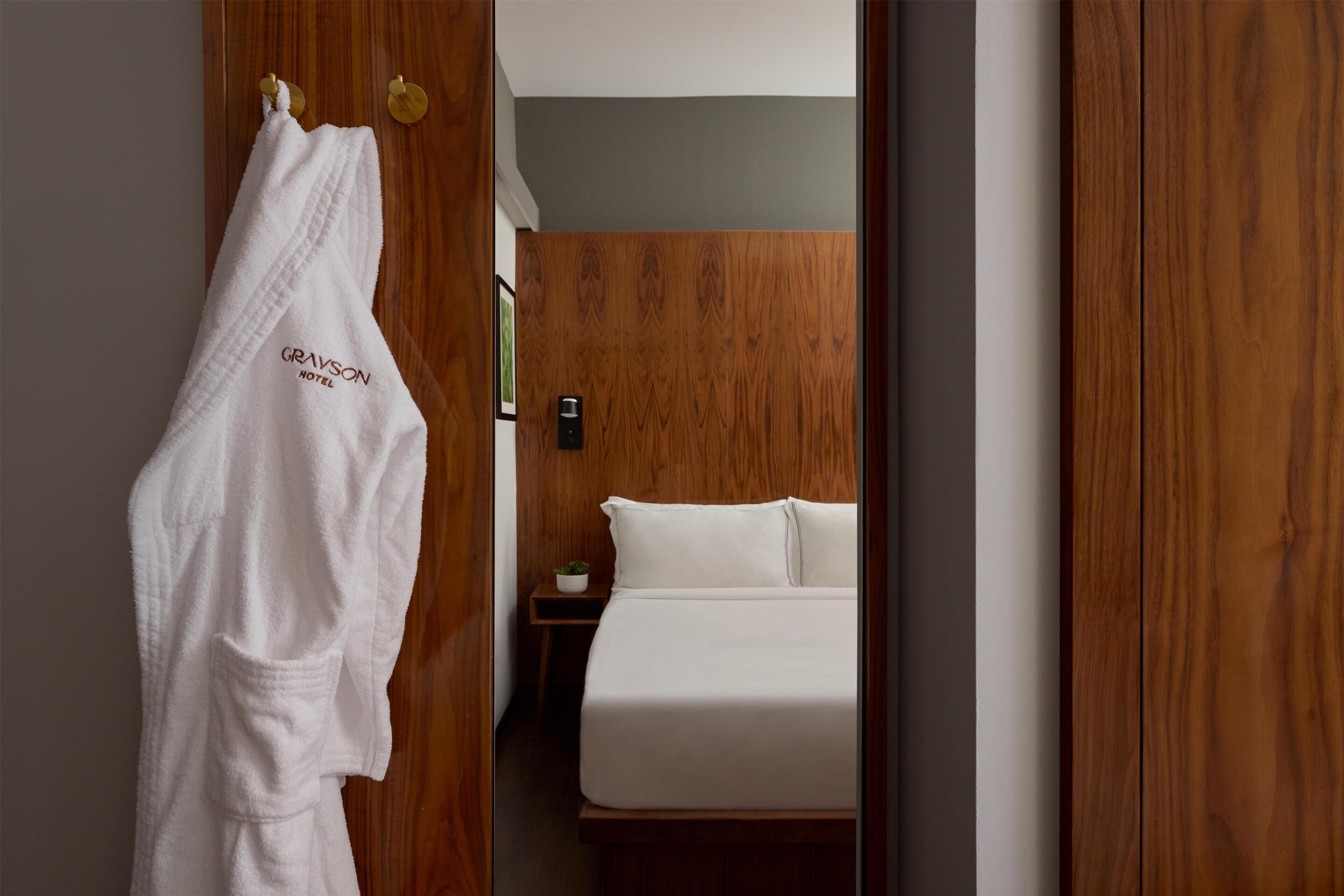 A hotel room with a wooden wardrobe, a white robe hanging on the door, and a bed with white bedding.