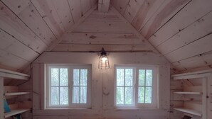 Um quarto de sótão acolhedor, com teto alto, vigas de madeira, duas janelas, uma cama com tapete estampado e estantes embutidas.