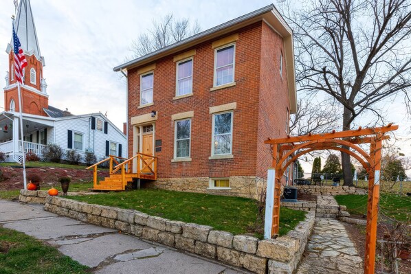 A two-story brick building with a stone foundation, a wooden porch, and a stone pathway leading to a wooden pergola.