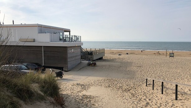 En byggnad vid stranden med en träfasad och en skylt med texten ”DES BOEKANIER STRANDKOLON”.