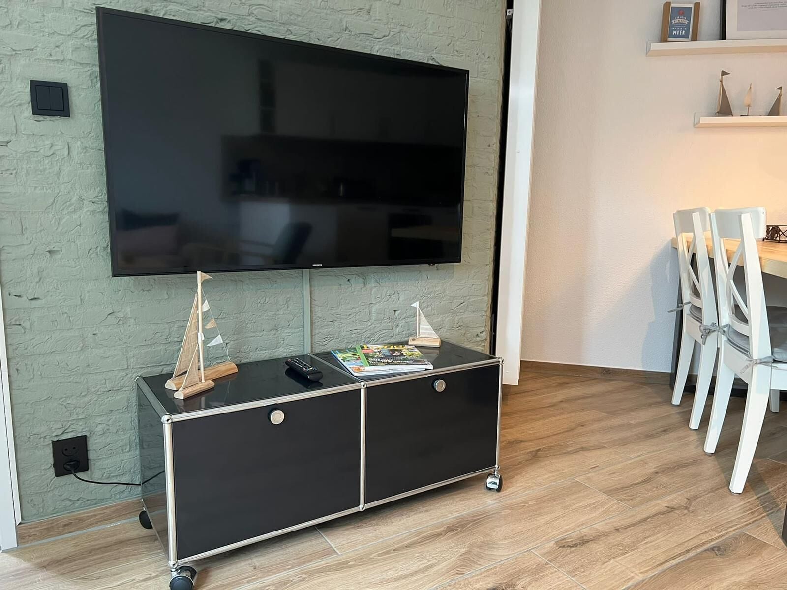 Ein modernes Wohnzimmer mit einem Flachbildfernseher an einer strukturierten Wand, einer schwarzen Unterhaltungsanlage inklusive Fernbedienung und einem Essbereich mit weißen Stühlen.