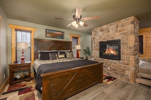 A bedroom with a stone fireplace, a bed with a wooden headboard, a ceiling fan, and a nightstand with a lamp.