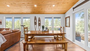 A wooden dining table with benches, a leather sofa, and large windows with a view of trees.