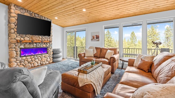 A cozy living room with a stone fireplace, leather sofas, and large windows offering a view of trees.