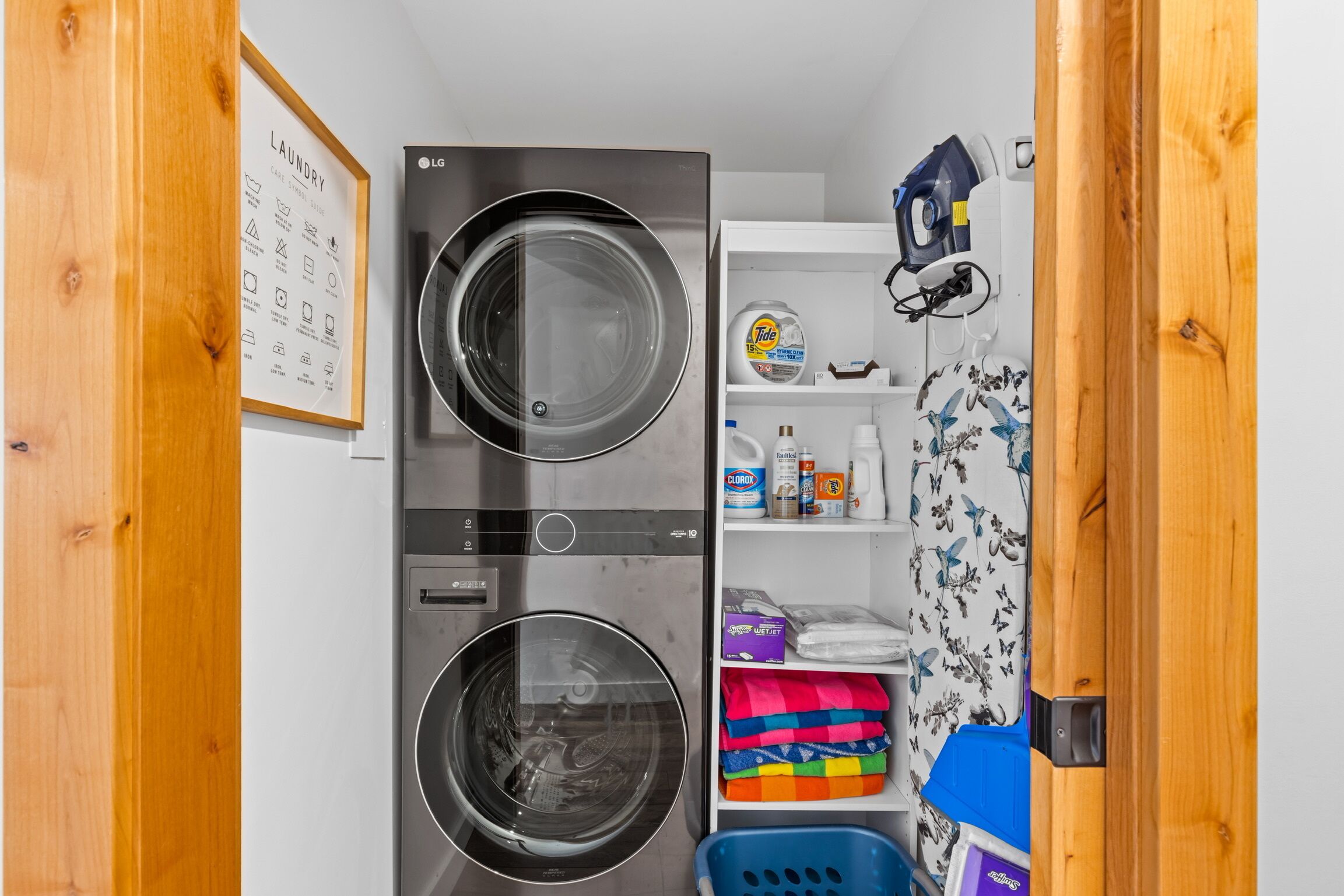Une buanderie équipée de deux machines à laver superposées, d’une étagère contenant des produits de lessive et d’un panier à linge bleu.