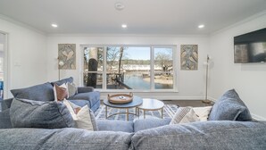 A living room with a gray sectional sofa, a round coffee table, and a large window with a view of a body of water and trees. - Cheerful & Stylish 4 Bedroom Lake-Side House (Hot Springs)