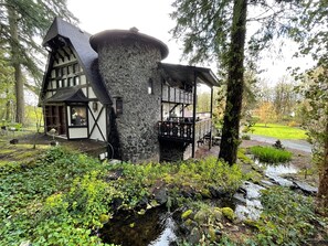 Une maison de campagne rustique, dotée d’une tour en pierre, de poutres en bois et d’un étang au premier plan.