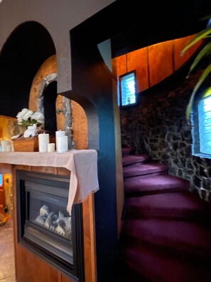 Una escalera de paredes de piedra con alfombra púrpura, una chimenea con repisa blanca y un ventanal con diseño decorativo.