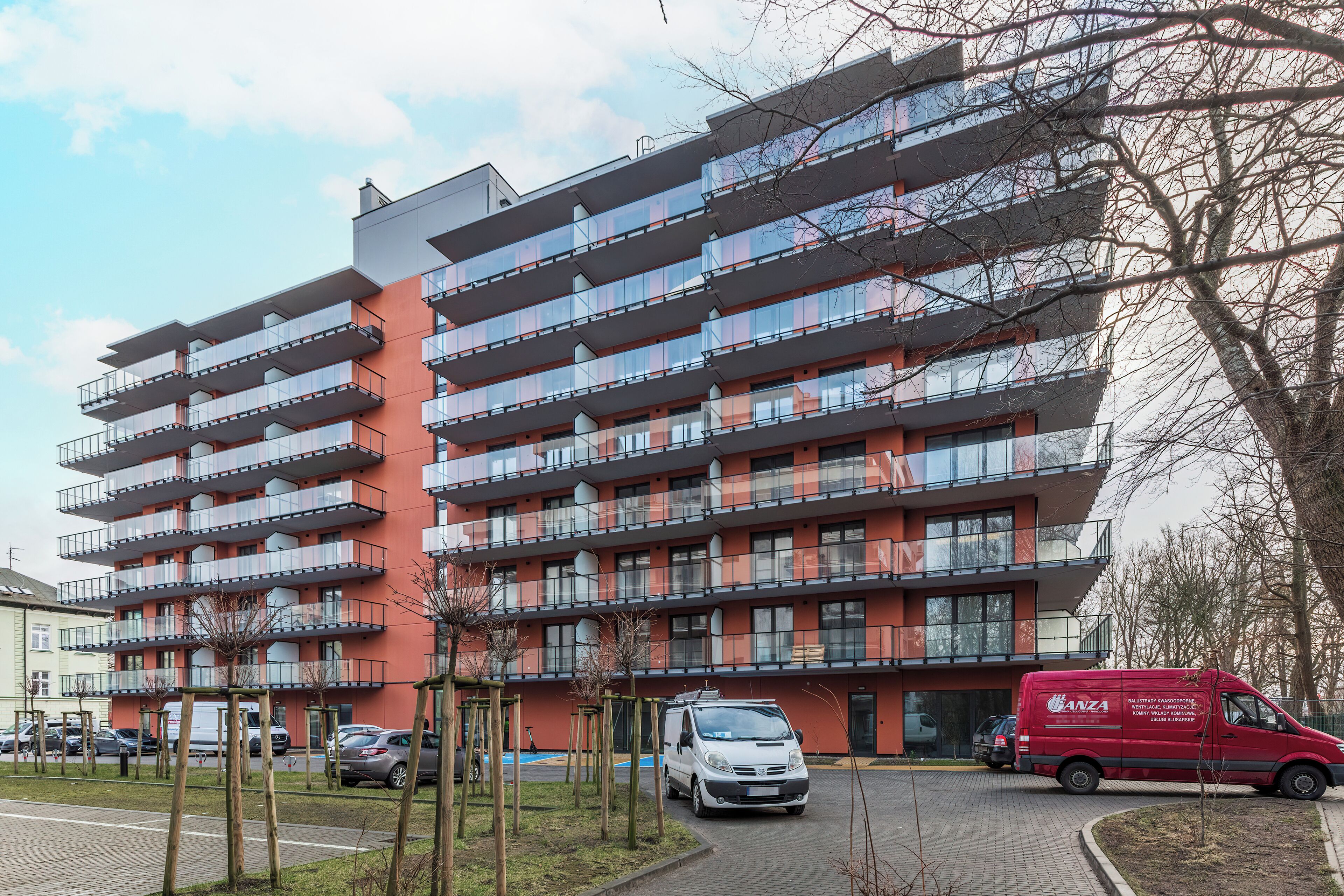 Un edificio residencial de varios pisos con balcones, un estacionamiento y una camioneta roja estacionada frente.