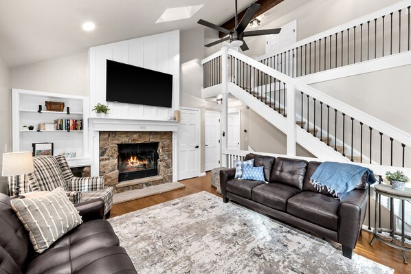 A living room with a fireplace, a staircase, a leather sofa, and a flat-screen TV. - Walk to RMNP, hot tub, mountain views and river access. (Estes Park)