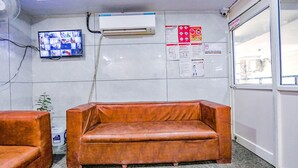 Ein Zimmer mit braunem Ledersofa, einem an der Wand montierten Fernseher, einer Klimaanlage und Informationsplakaten.