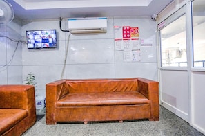 A room with a brown leather sofa, a mounted TV, air conditioning unit, and informational posters on the wall. - Fabexpress Rama (New Delhi)