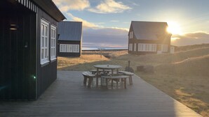A wooden deck with a round table and benches, leading to a beach with two houses in the background.