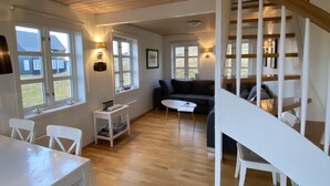A spacious living area with a wooden floor, white furniture, a staircase, and large windows.
