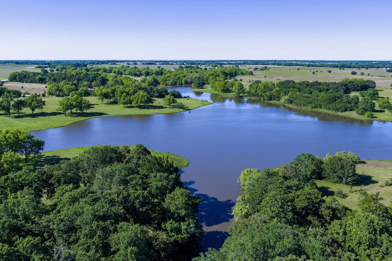 A large body of water surrounded by dense green trees and open fields.