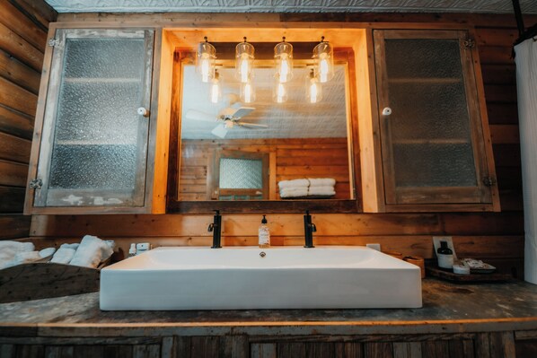 A wooden cabin bathroom with a large white sink, wooden cabinets, and a mirror with frosted glass panels.