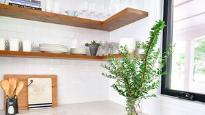 Una cocina con armarios blancos, encimera de mármol, repisa de madera con vajilla y una planta en maceta.