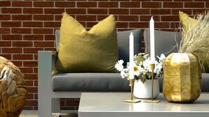 Un área de estar moderna al aire libre con un sofá gris, una mesa de centro blanca y detalles decorativos como un jarrón con flores y una vela blanca alta.