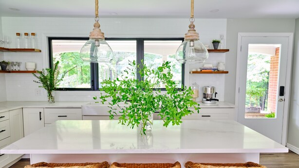 Una cocina moderna con una isla grande, luces colgantes, taburetes de mimbre y vistas al exterior con vegetación.