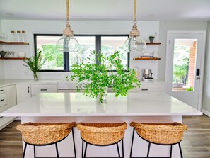 Een moderne keuken met een groot schiereiland, hangende kroonluchters, rieten barstoelen en uitzicht op groen.