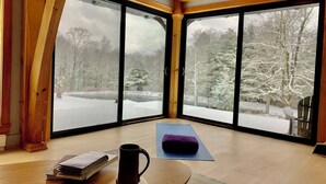 Una stanza con ampie finestre, pavimento in legno, un tappetino per yoga, una pila di libri e una tazza.