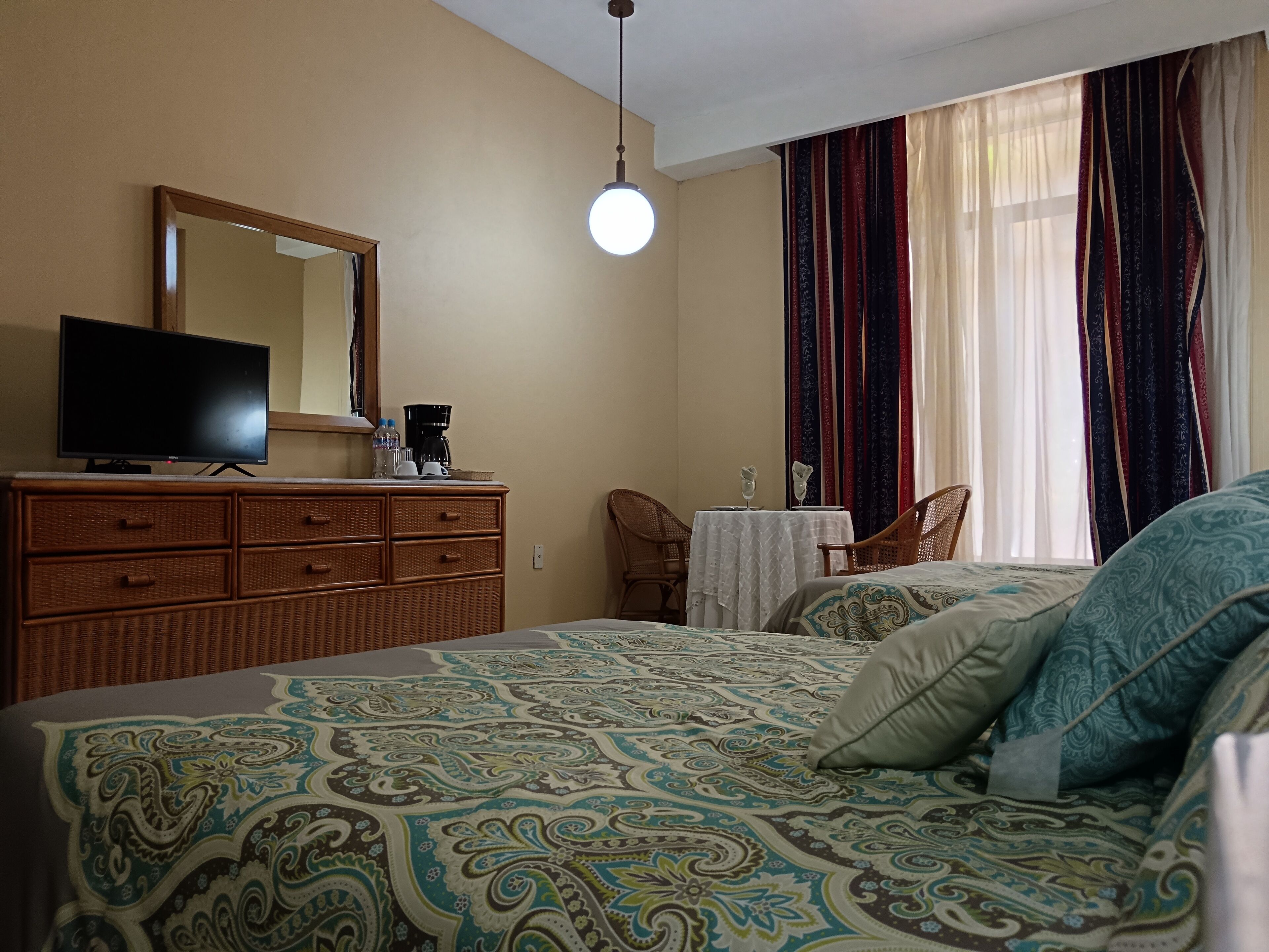 Un dormitorio con cama, cómoda, televisor, mesa con silla y ventana con cortinas.