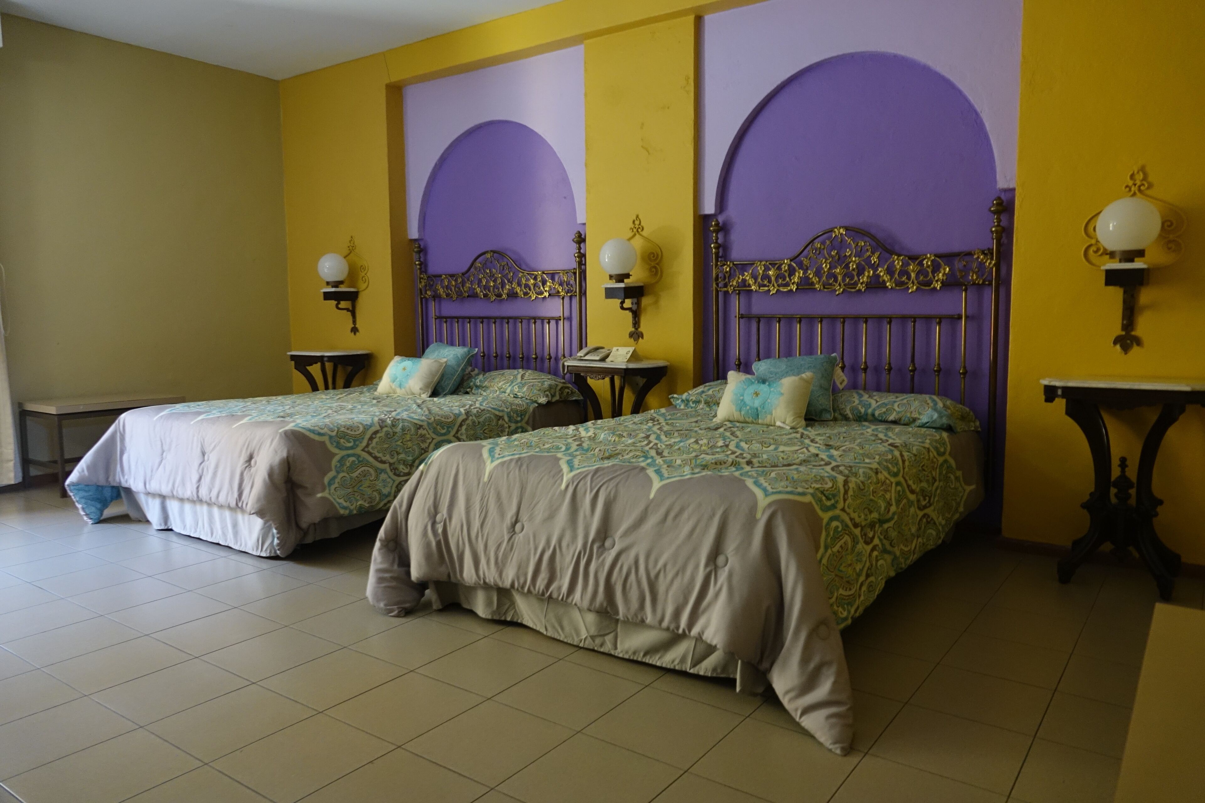 Habitación con dos camas, cada una con ropa de cama estampada y cabeceros, una mesita pequeña y lámparas de pared.