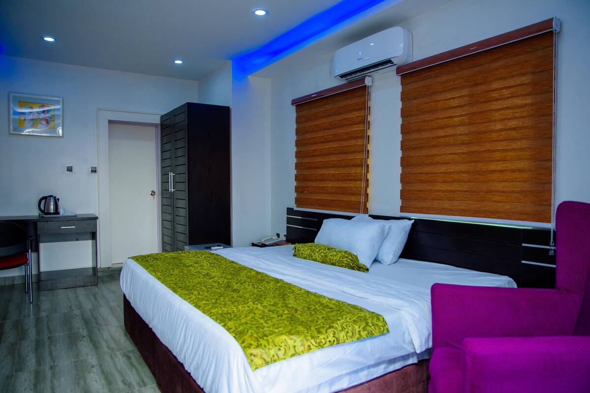 A hotel room with a bed, a desk, a chair, and an air conditioning unit.
