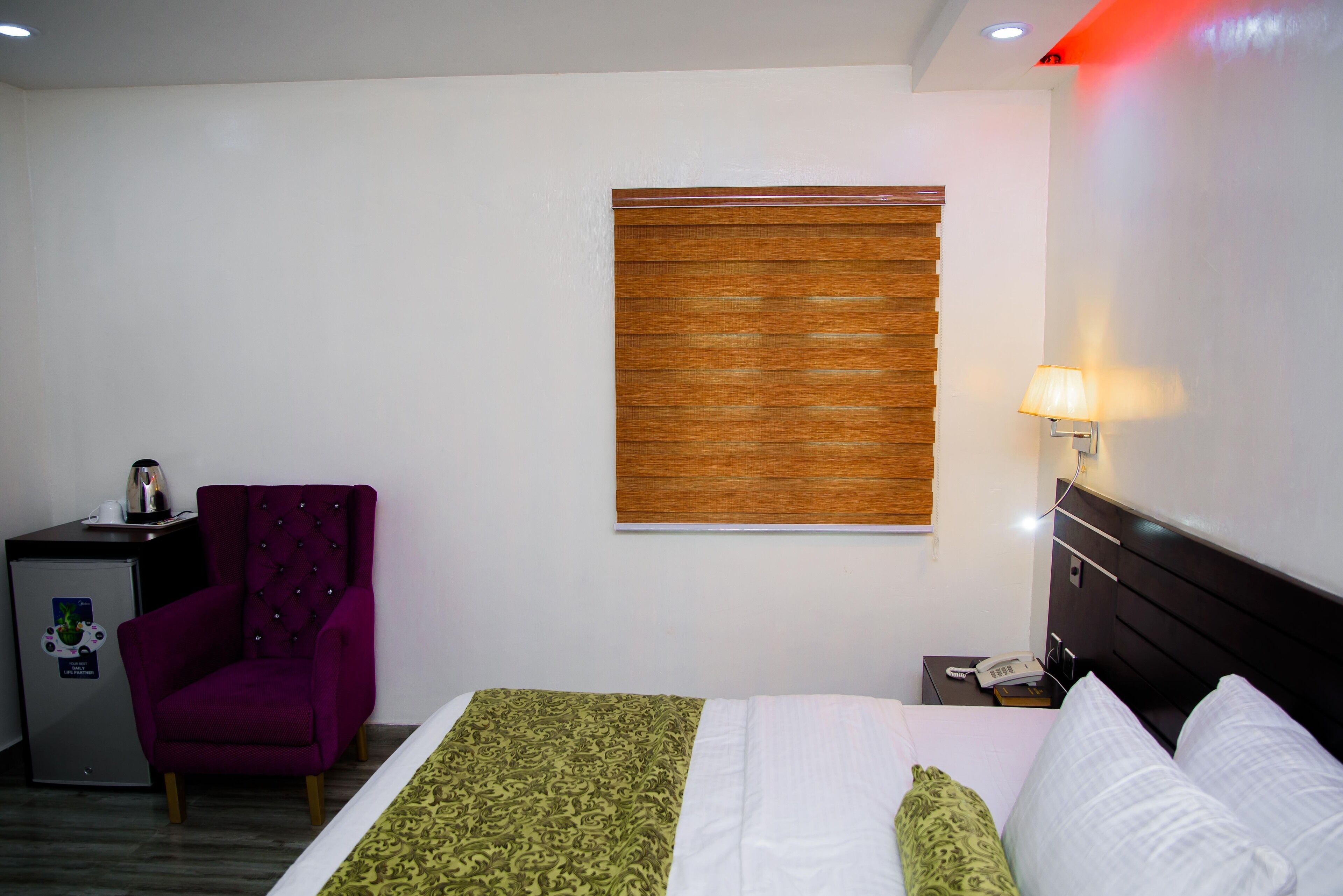 A hotel room with a bed, a purple armchair, a small desk with a kettle, a nightstand with a lamp, and a window with blinds.