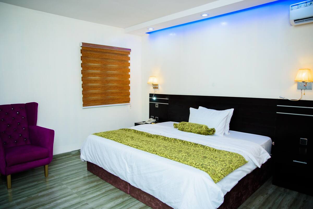 A hotel room with a bed, a chair, a nightstand, and a wall-mounted lamp.