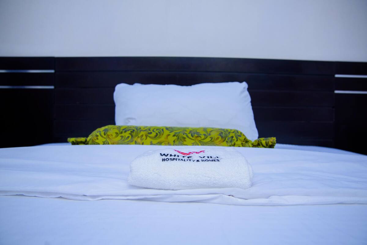 A neatly made bed with white linens, a green patterned pillow, and a white pillow. A towel with the text "WHITE VILL HOSPITALITY & HOMES" is placed on the bed.