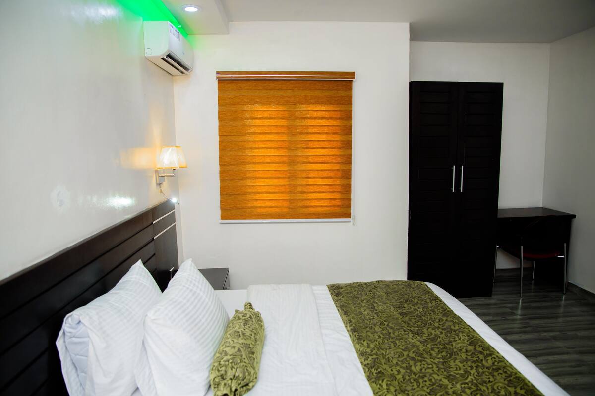 A hotel room with a bed, a desk, a wardrobe, and a window with blinds.