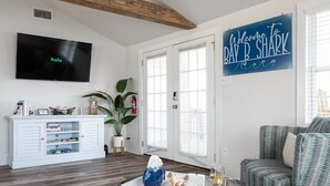 A living room with a flat-screen TV displaying Hulu, a white entertainment unit, a glass coffee table with decorative items, a blue chair, and a 'Welcome to Bay B. Shark' sign.