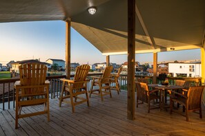 Une terrasse en bois, agrémentée de chaises et de tables, qui offre une vue sur une étendue d’eau et des bâtiments.