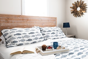 A neatly made bed with a white and blue fish-patterned duvet, a tray with a muffin, berries, and a glass of blue liquid, and a wooden headboard.
